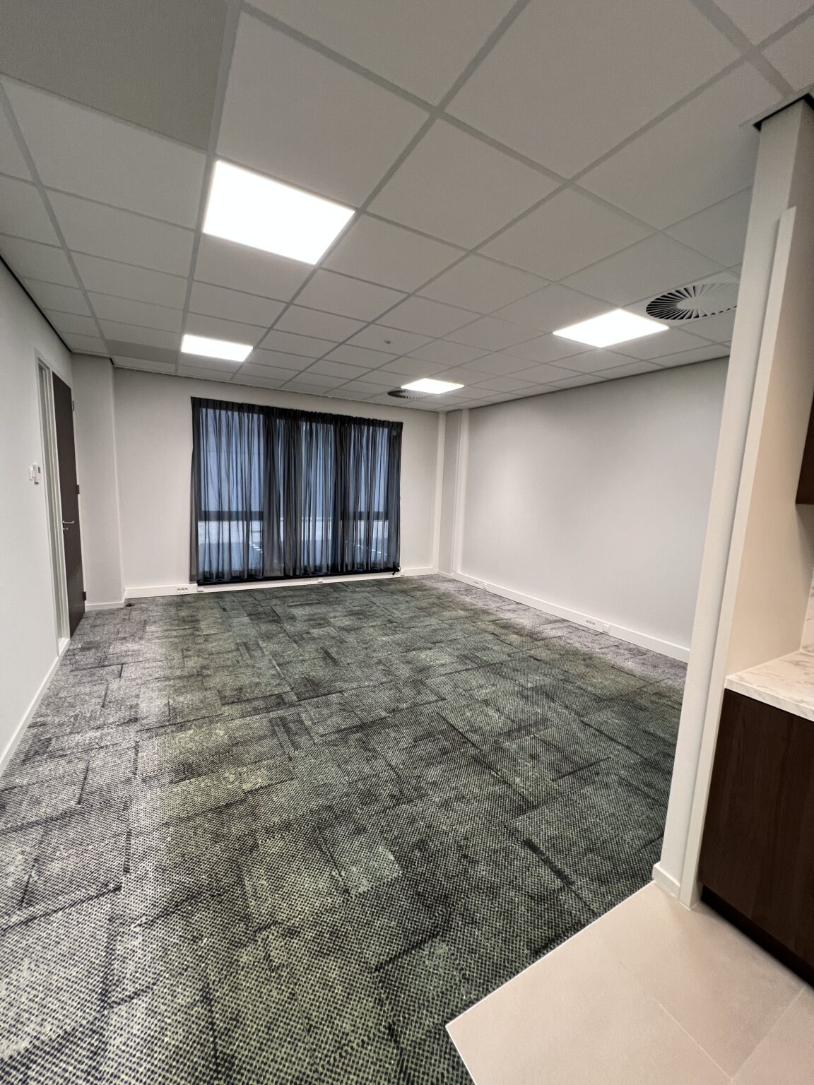 Leeg kantoorpand met tapijt en grote ramen, verlicht door inbouw plafondlampen.