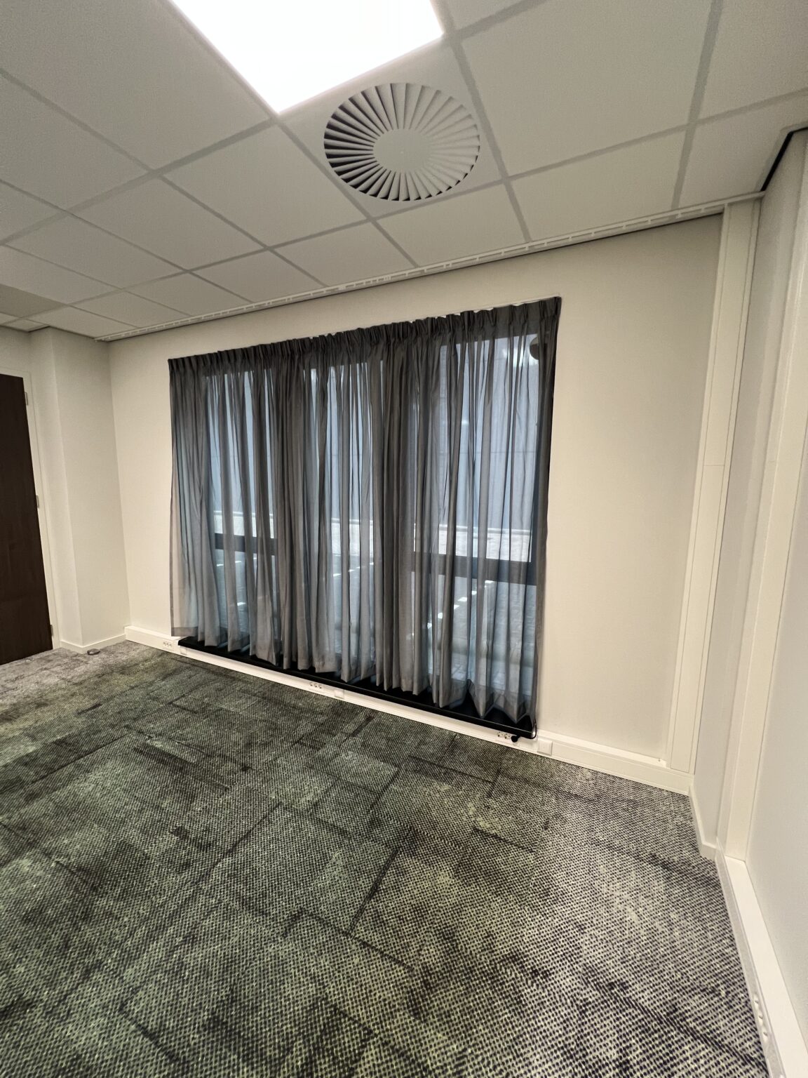 Een kantoorkamer met een grijs tapijt, gesloten gordijnen en een systeemplafond met ventilatierooster.